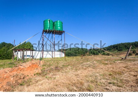 Forest Farm with two Large green colored water tanks on tall steel stand tower above round reservoir below in open field in blue sky landscape.