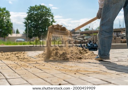 Man working with a shovel picks up sand from a large pile to scatter it through the cracks between the paving slabs. Laying paving slabs on a sunny summer day