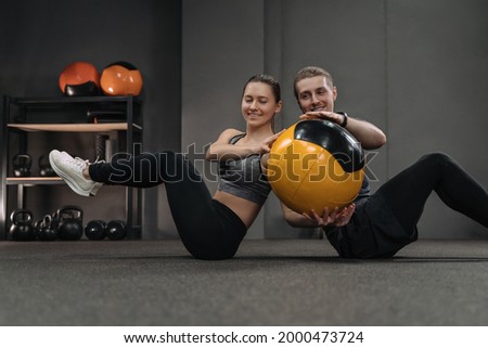 Young couple doing abs exercises with medicine ball at dark crossfit gym. Couple sitting back to back on floor and doing workout by passing ball to each other. Sporty woman and man training together Royalty-Free Stock Photo #2000473724