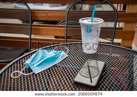 Concept of pandemic travel, morning drink, without mask when drinking coffee in public place, opening of the cafe after lockdown. cold drink, face mask on metal table.