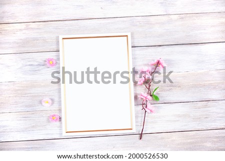 A white and gold photo frame lies on a board table with pink flowers. Festive photo frame mockup.