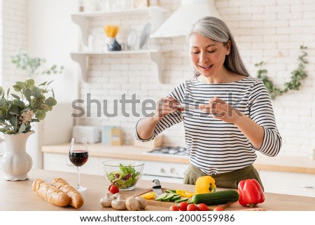 Happy mature middle-aged woman wife mother blogger taking photo of her cooking process while preparing vegetable salad vegetarian food meal in the kitchen at home. Food blogging