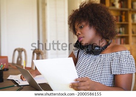 African american woman working in living room using laptop, paperwork and talking on smartphone. technology and communication, flexible working from home.