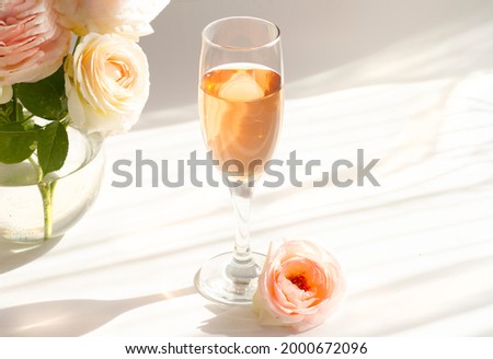 A glass of semi-sweet pink wine Pinot rose . Next to a bouquet of antique terry roses Pierre de Ronsard, Eden roses. Striped shadows on a white background.
