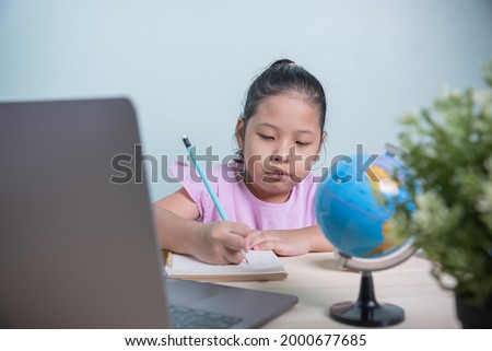 Asian kid girl writing follows teacher for learning an online lesson by video call the Internet from laptops in the room. The spread of the coronavirus has forced students to study online from home.