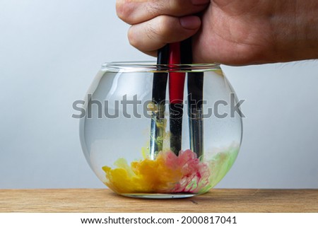 Paint brushes dipped in a jar of water on a white background. The paint spreads in the water.