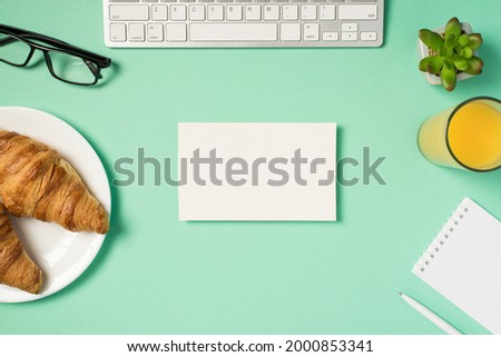 Top view photo of workplace white rectangular card in the middle keyboard flowerpot glasses copybook pen plate with croissants and glass of juice on isolated turquoise background with blank space