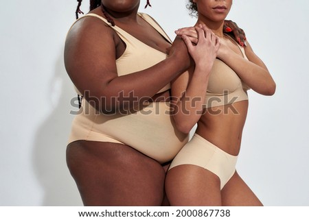 Cropped shot of bodies of two african american women in beige underwear with different weight supporting each other, posing together isolated over gray background. Friendship, body positive