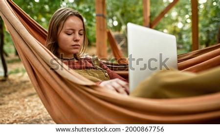 Top view of a young girl surfing the net on the fresh air while chilling in the hammock, wearing brown jacket and a beige overall, holding laptop on her knees