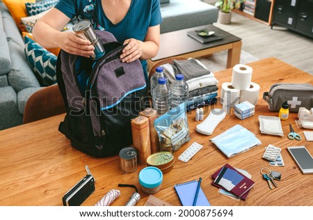 Unrecognizable woman putting cans of food to prepare emergency backpack in living room Royalty-Free Stock Photo #2000875694
