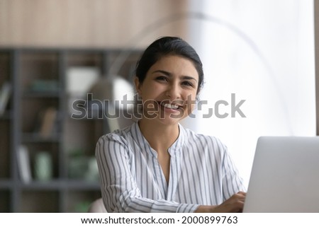White collar. Happy young indian female accountant consultant sit at workplace by laptop take break in typing document look at camera. Headshot portrait of cheerful ethnic woman use pc in office work Royalty-Free Stock Photo #2000899763