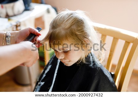 Adorable toddler girl getting his first haircut. Happy cute child sitting in domestic kitchen. Mother making a hair style to cute little baby girl daughter. During corona virus pandemic.