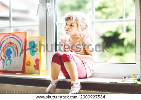 Cute little toddler girl by window with rainbow painted with colorful colors. Preschool on bad weather rainy day looking out the window. Alone upset kid