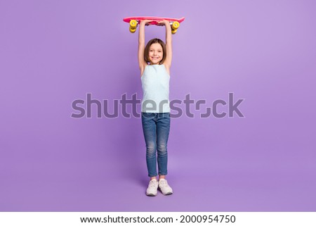 Full body photo of attractive happy sportive little girl raise longboard above head isolated on purple color background