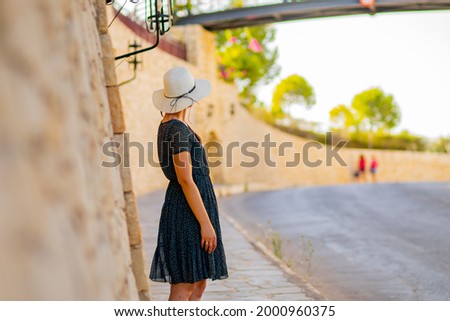 A young woman is on the sidewalk in the beautiful Cretan town of Hersonissos.
She is wearing a white hat and a blue dress. In the distance, other tourists approach by the high stone wall