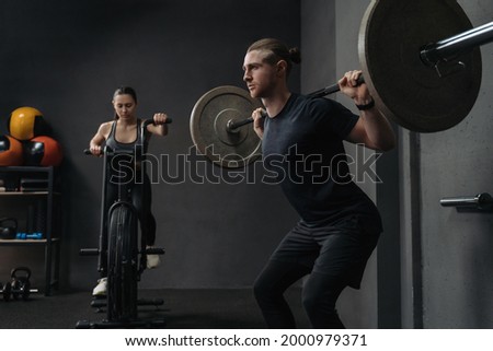 Couple have crossfit training at gym. Attractive fit woman is working out on exercise bike, while muscular man training with barbell. Functional and circuit training concept Royalty-Free Stock Photo #2000979371