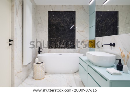 Elegant bathroom with black and white marble tiles on floor and walls, big freestanding bathtub and blue chest of drawers under washbasin Royalty-Free Stock Photo #2000987615