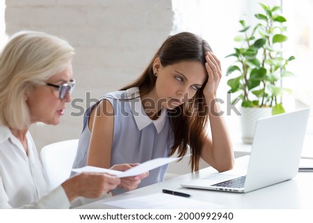 Unhappy young female employee bored with middle-aged colleague discuss paper document at meeting. Tired woman worker bothered annoyed by senior coworker lecture about paperwork at team briefing. Royalty-Free Stock Photo #2000992940
