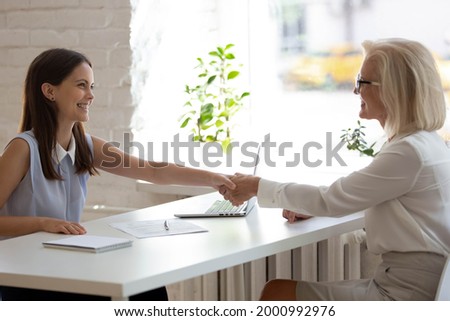 Smiling young Caucasian recruiter shake hand get acquainted greet with senior female client or customer in office. Happy businesswoman handshake close deal make agreement with middle-aged employee.