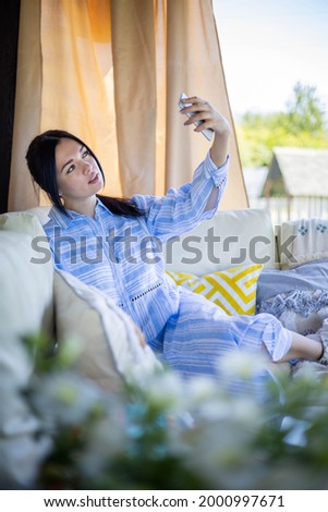 Beautiful young mixed race brunette woman relaxing at outdoor summer terrace posing taking selfie use smartphone. Adorable Asian female sitting on comfortable couch photographing on mobile phone