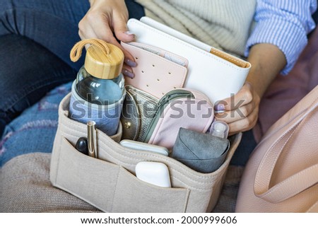 Closeup woman hands putting personal accessories into felt organizer. Concept of storage organization in female bag. Modern keeping system in handbag with tablet, passport, bottle water, cosmetics Royalty-Free Stock Photo #2000999606
