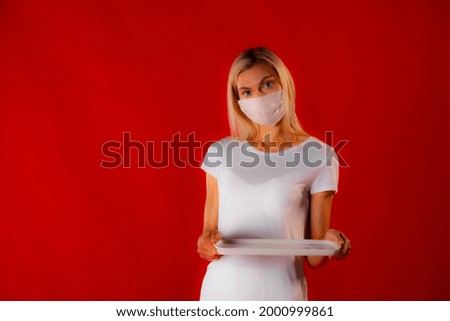 Copy space closeup of white dressed girl looking on tray. Copy space photo of slim blonde in white medical mask and white outfit on matte red background holding mockup plate