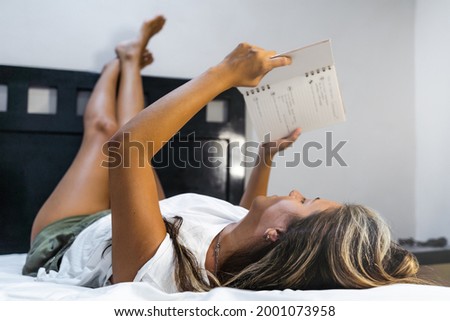 young hispanic woman lying in bed in pajamas with legs raised reading notes in her personal diary
