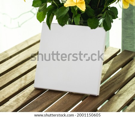 White square empty printable canvas stands on a table with a vase of flowers. Mockup canvas (947)
