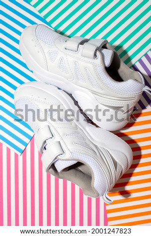 vertical top view two white kid sneakers with velcro fasteners for comfortable footwear on an abstract geometric paper multicolored striped background with a hard light.