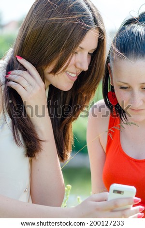 2 beautiful young women best girlfriends having fun looking at screen one showing another message on white mobile cell phone happy smile on summer green outdoors copy space background portrait picture