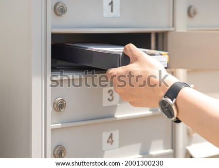Mail man delivering small parcel in mailbox by house number, selective focus. Person’s hand holding package put in metal storage. People get packet from opened cabinet door. Sender, receiver concept.