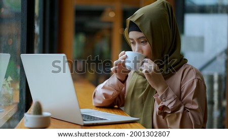 Portrait young pretty muslim woman works with laptop computer in coffee shop with happiness, she hold a coffee cup to drink while looking at screen, muslim business working concept
