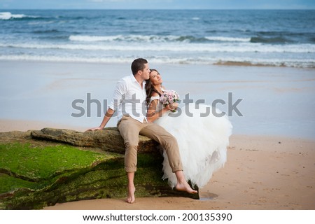 Young beautiful bride in a wedding dress and a handsome groom sitting on a picturesque rock at the beach and looking at the ocean