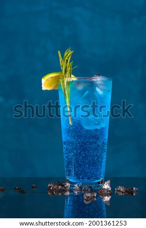 Fresh BLUE LAGOON summer cocktail on curacao background, with melted ice around it, garnished with lime, rosemary and cranberries.