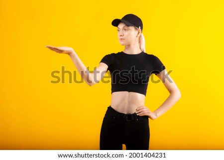 Thin girl in black clothes holding right hand up mockup space. Pretty blond blue-eyed girl model in black high waist jeans, black short t-shirt and black cap posing on yellow matte background