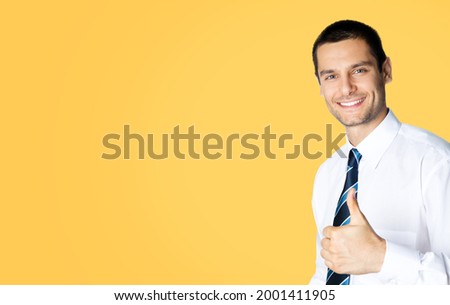 Portrait of smiling businessman in white shirt and tie, showing thumb up like hand sign gesture, isolated over yellow colour background. Happy confident business man gesturing. Copy space area. Male.