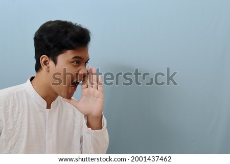 Portrait of angry Asian man in koko shirt or white muslim shirt standing against blue background, shouting loud with arm at his face Royalty-Free Stock Photo #2001437462