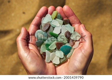 A person holds a collection of multi colored sea glass in their cupped hands with the sand in the background Royalty-Free Stock Photo #2001439790