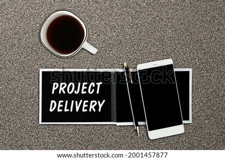 A notebook with black pages, smartphone and a cup of coffee on a grey background. The inscription PROJECT DELIVERY on the notepad.