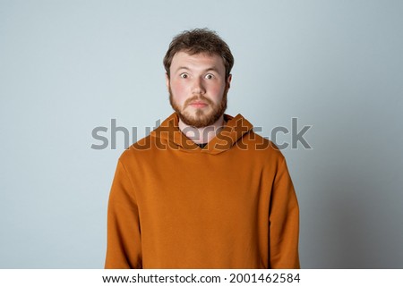 Portrait of shocked and scared bearded modern young man, looking amazed at something embarrassing, standing in stylish hoodie against studio gray background