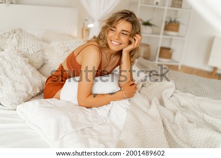 Smiling  young woman stretching in bed waking up in cozy bedroom with bohemian interior style. Female wearing in comfort pajamas enjoying early morning,.