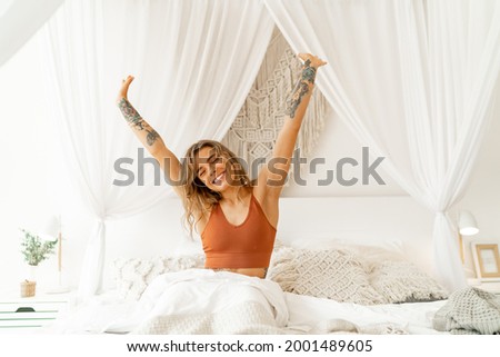 Smiling  young woman stretching in bed waking up in cozy bedroom with bohemian interior style. Female wearing in comfort pajamas enjoying early morning,. Royalty-Free Stock Photo #2001489605