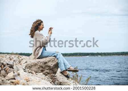 Young woman sitting on rocky bank taking photo of landscape using smartphone.