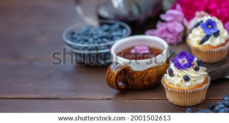 Cupcake with cream decorated with violet flowers lying next to a mug of tea among the flowers of peonies and scattered honeysuckle berries