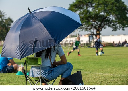 Parents sitting and watching their son playing football in a school tournament on a clear sky and sunny day. Sport, outdoor activity with lifestyle and a happy family. Soccer mom and dad concept. Royalty-Free Stock Photo #2001601664