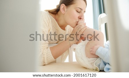 Happy smiling mother kissing feet of her little baby lying in cradle. Concept of parenting, family happiness and baby development.