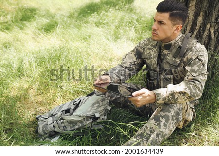 Soldier with backpack using tablet near tree in forest