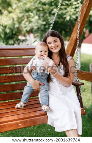 Happy beautiful woman, young mother playing with her adorable baby son and sitting on wooden swing, cute little boy, enjoying together a sunny warm day playing on the lawn in a summer garden.