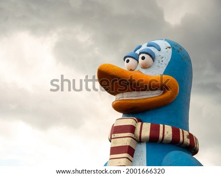 Sculpture of blue color, comic, smiling, optimistic penguin with big eyes and striped scarf. Head on overcast sky background.