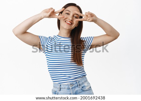 Beautiful carefree girl enjoying day, showing peace v-sign, disco fingers near eyes, smiling happy and satisfied, enjoy summer, standing against white background